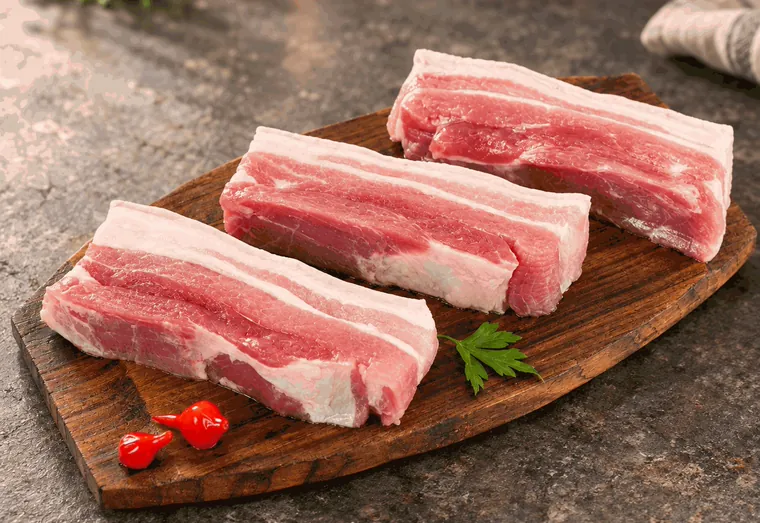 Três pedaços de panceta crua, com as camadas de carne e gordura, sobre tábua.