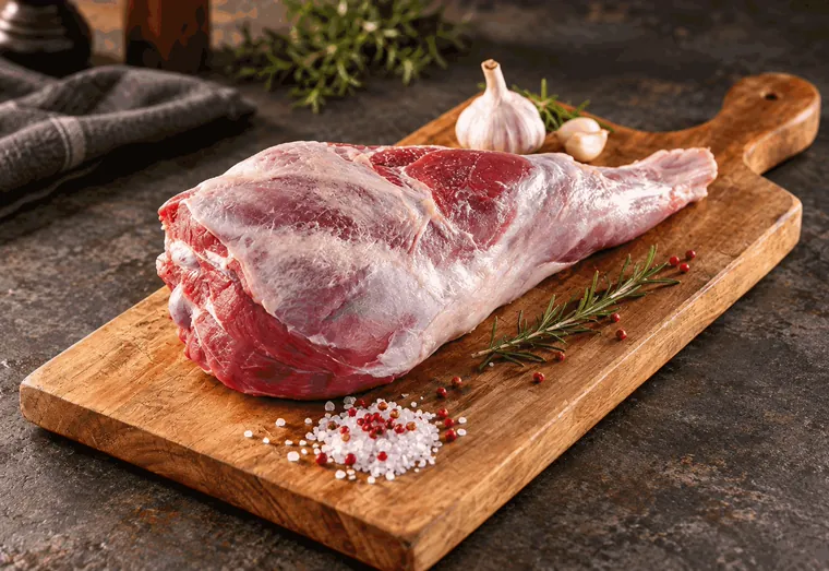Pernil de cordeiro cru sobre tábua de madeira, com alho, alecrim e sal grosso.