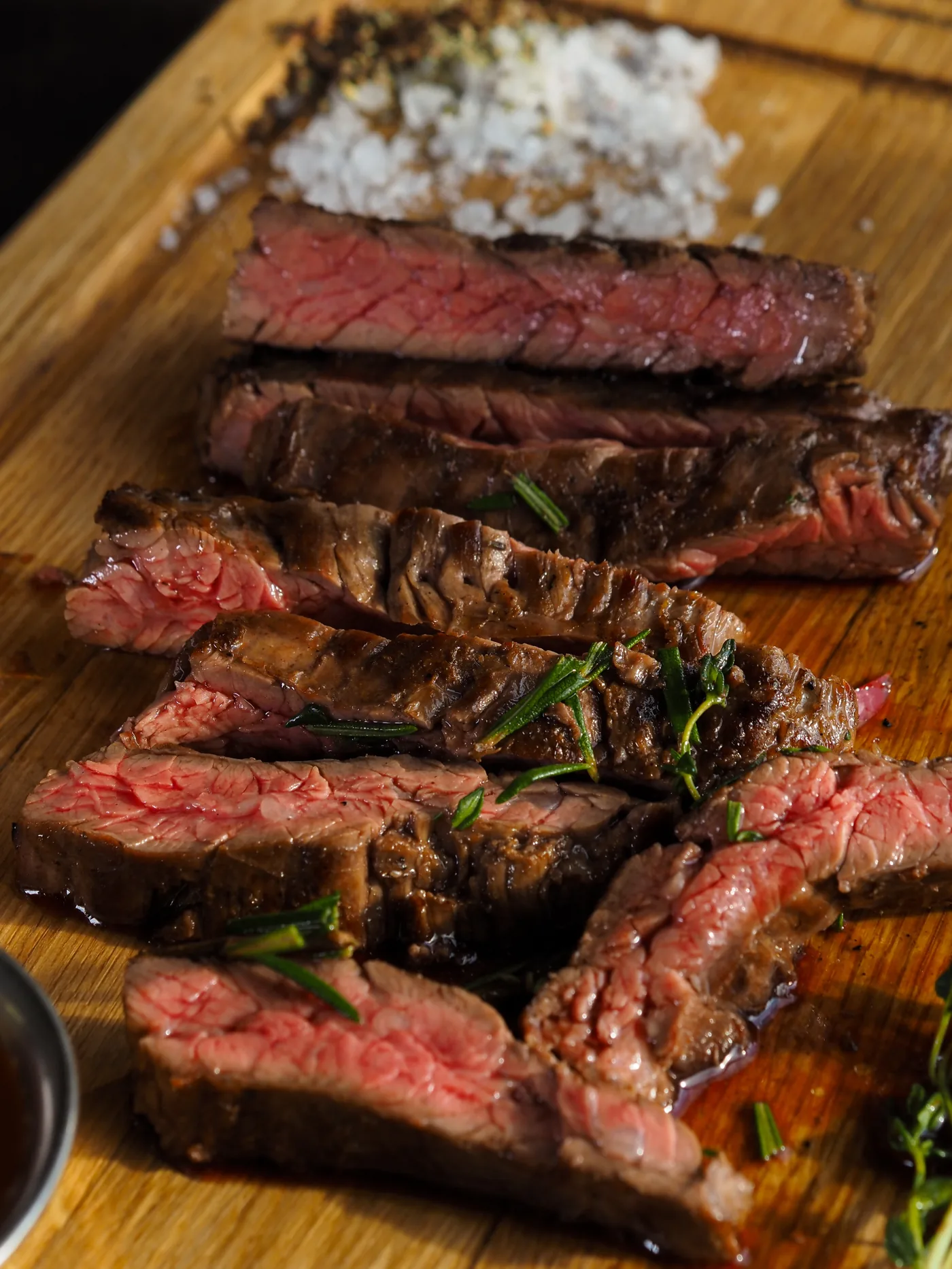 Peça de carne bovina fatiada sobre tábua de madeira, com sal grosso e alecrim.